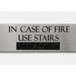 A rectangular brushed-metal emergency exit sign mounted on a textured white wall. The sign features the text 'IN CASE OF FIRE USE STAIRS' in black capital letters above a black rectangular panel containing raised Braille lettering.