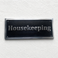 A rectangular black housekeeping sign with silver raised lettering and a metallic silver border, mounted on a textured white wall.