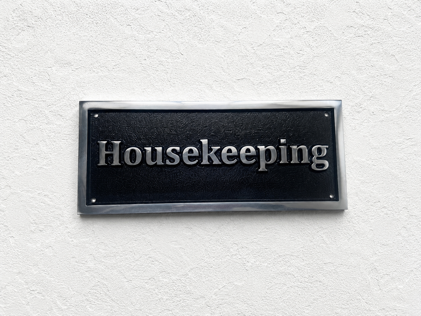 A rectangular black housekeeping sign with silver raised lettering and a metallic silver border, mounted on a textured white wall.