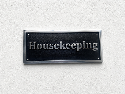 A rectangular black housekeeping sign with silver raised lettering and a metallic silver border, mounted on a textured white wall.