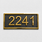A rectangular house number plaque featuring the gold numerals '2241' on a textured black background, bordered by a polished gold frame, mounted on a light gray textured wall.