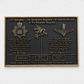 A rectangular black commemorative plaque with gold-toned metal border, raised lettering, and military insignia mounted on a white textured wall.