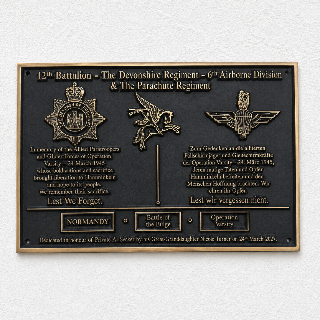 A rectangular black commemorative plaque with gold-toned metal border, raised lettering, and military insignia mounted on a white textured wall.