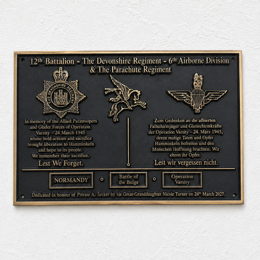 A rectangular black commemorative plaque with gold-toned metal border, raised lettering, and military insignia mounted on a white textured wall.