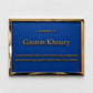 A rectangular blue memorial plaque with a gold metallic border, mounted on a textured white wall. The plaque features raised gold text reading 'In memory of Gaston Khoury' and a shorter commemorative paragraph below.