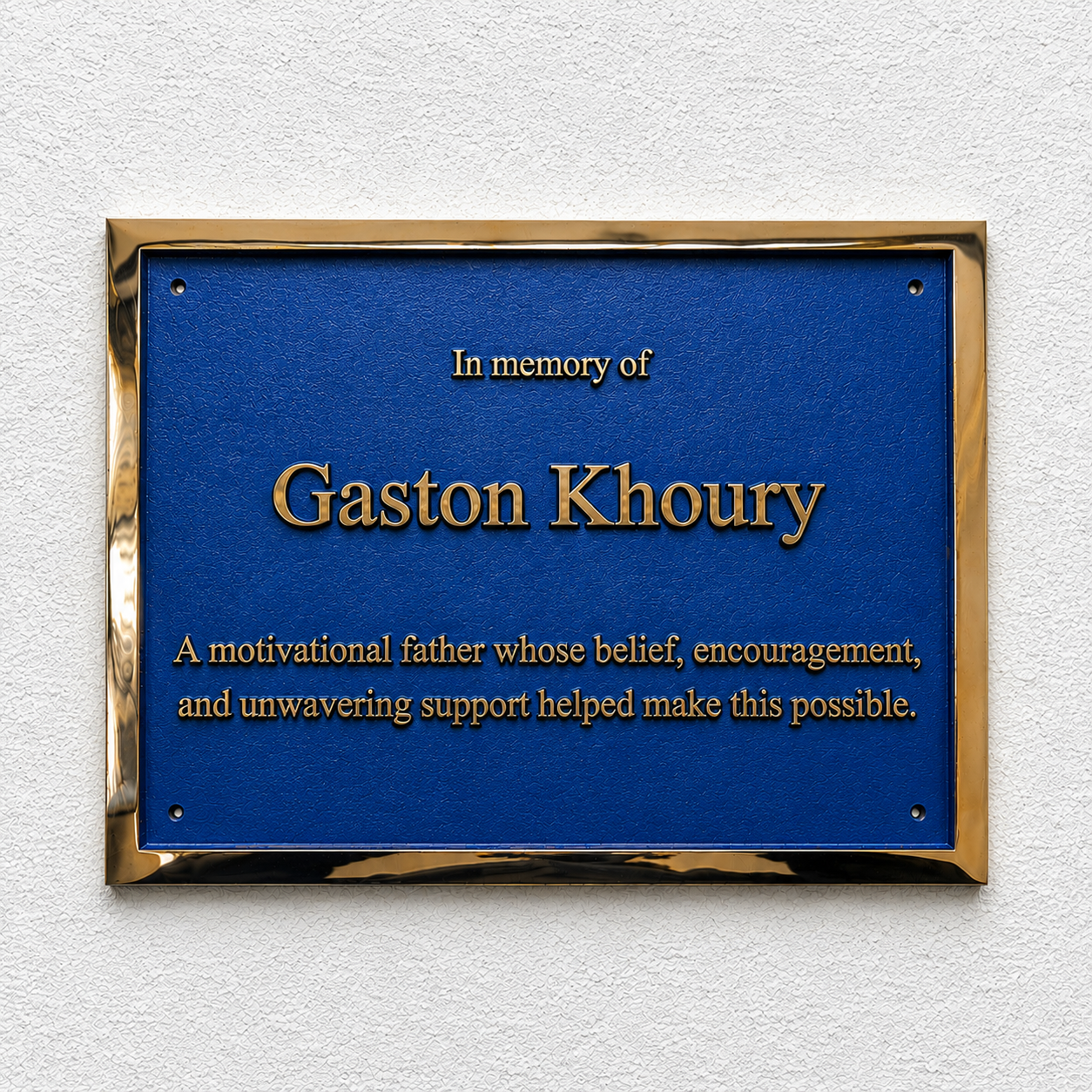 A rectangular blue memorial plaque with a gold metallic border, mounted on a textured white wall. The plaque features raised gold text reading 'In memory of Gaston Khoury' and a shorter commemorative paragraph below.