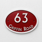 An oval house number plaque reading '63 Griffin Road' in raised white lettering on a textured red background, mounted against a white wall.