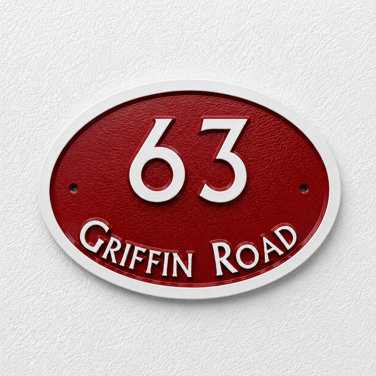 An oval house number plaque reading '63 Griffin Road' in raised white lettering on a textured red background, mounted against a white wall.