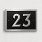 A rectangular house number plaque with the number 23 in polished silver-tone metal over a textured black background, mounted on a white textured wall.