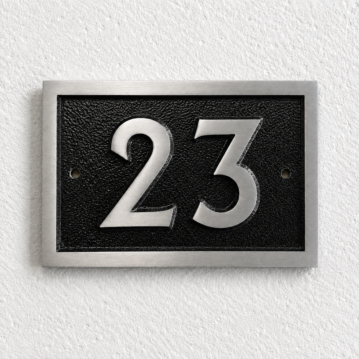A rectangular house number plaque with the number 23 in polished silver-tone metal over a textured black background, mounted on a white textured wall.