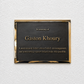 A rectangular black memorial plaque with a polished gold-toned border, mounted on a textured white wall. The plaque reads 'In memory of Gaston Khoury' with additional commemorative text below.