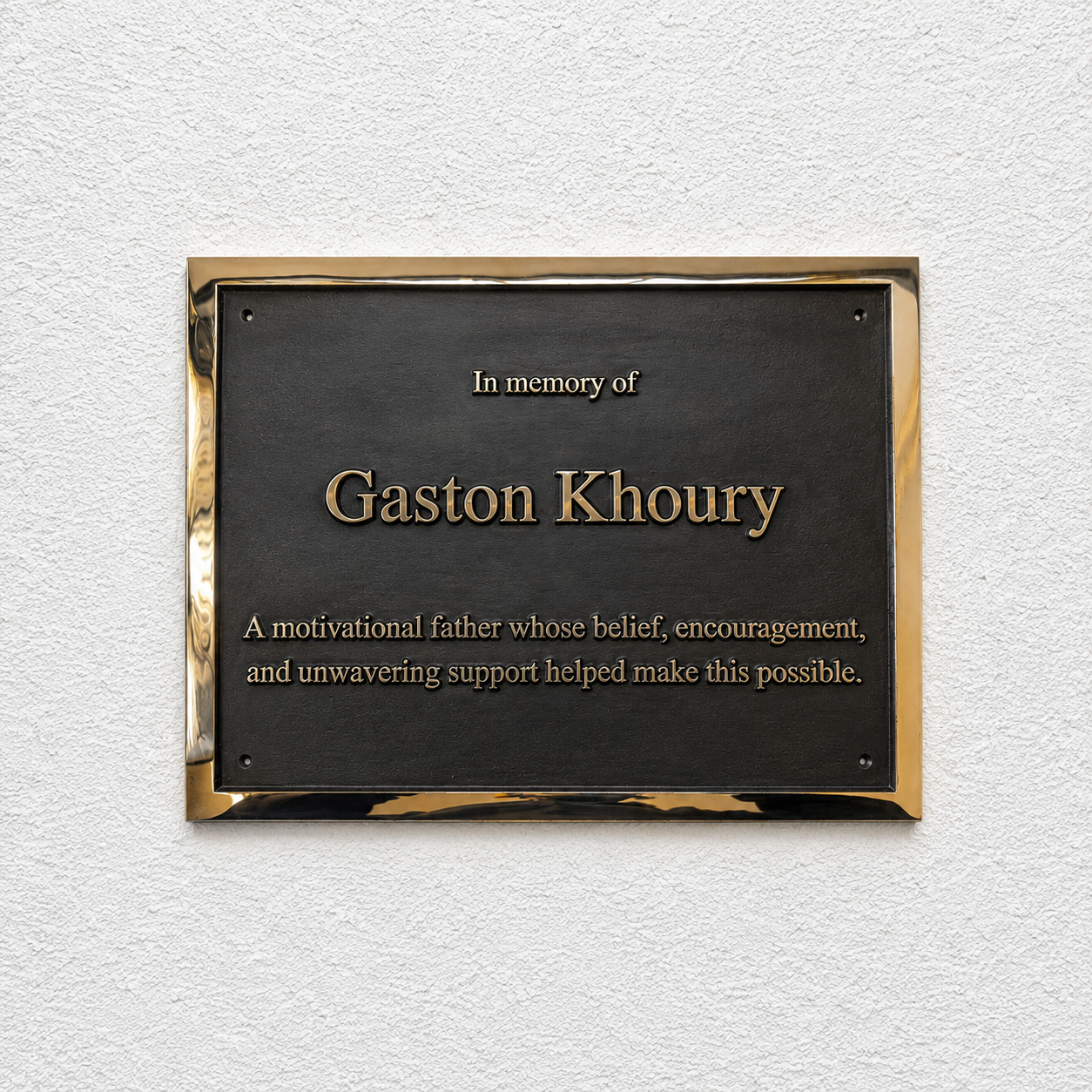 A rectangular black memorial plaque with a polished gold-toned border, mounted on a textured white wall. The plaque reads 'In memory of Gaston Khoury' with additional commemorative text below.