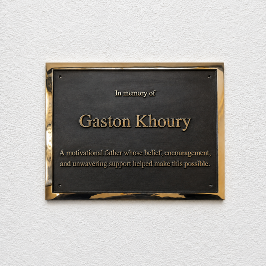 A rectangular black memorial plaque with a polished gold-toned border, mounted on a textured white wall. The plaque reads 'In memory of Gaston Khoury' with additional commemorative text below.