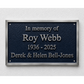 A rectangular dark blue memorial plaque with a brushed silver metal border, mounted on a light textured wall. The plaque features raised silver lettering that reads 'In memory of Roy Webb 1936 - 2025 Derek & Helen Bell-Jones'.