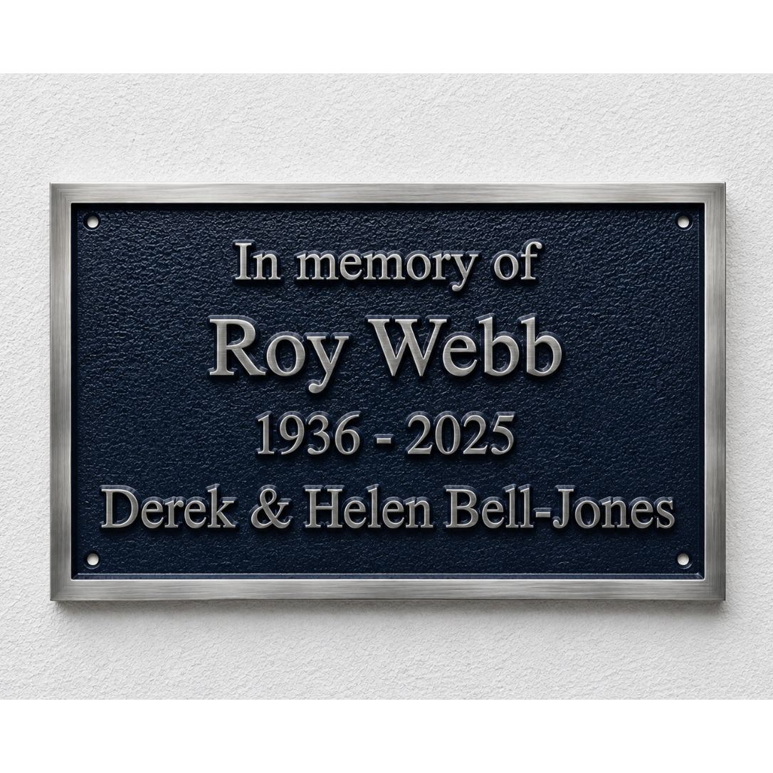 A rectangular dark blue memorial plaque with a brushed silver metal border, mounted on a light textured wall. The plaque features raised silver lettering that reads 'In memory of Roy Webb 1936 - 2025 Derek & Helen Bell-Jones'.