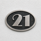 An oval house number plaque featuring the numbers '21' in brushed silver, mounted on a textured white stucco wall.