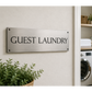 A brushed metal sign reading 'GUEST LAUNDRY' mounted on a light beige wall, with a doorway opening to a clean laundry room containing a washing machine and dryer, folded white towels, shelves, and plants.