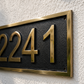 A close-up of a house number plaque showing the numbers 2241 in raised gold lettering on a textured black background, mounted on a white stucco wall.
