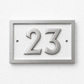 A rectangular house number plaque displaying '23' in raised metallic numerals on a textured light background, mounted on a white wall.