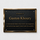 A rectangular black memorial plaque with a polished gold-toned border, mounted on a light textured wall. The plaque reads 'In memory of Gaston Khoury' and includes a short tribute below in gold lettering.