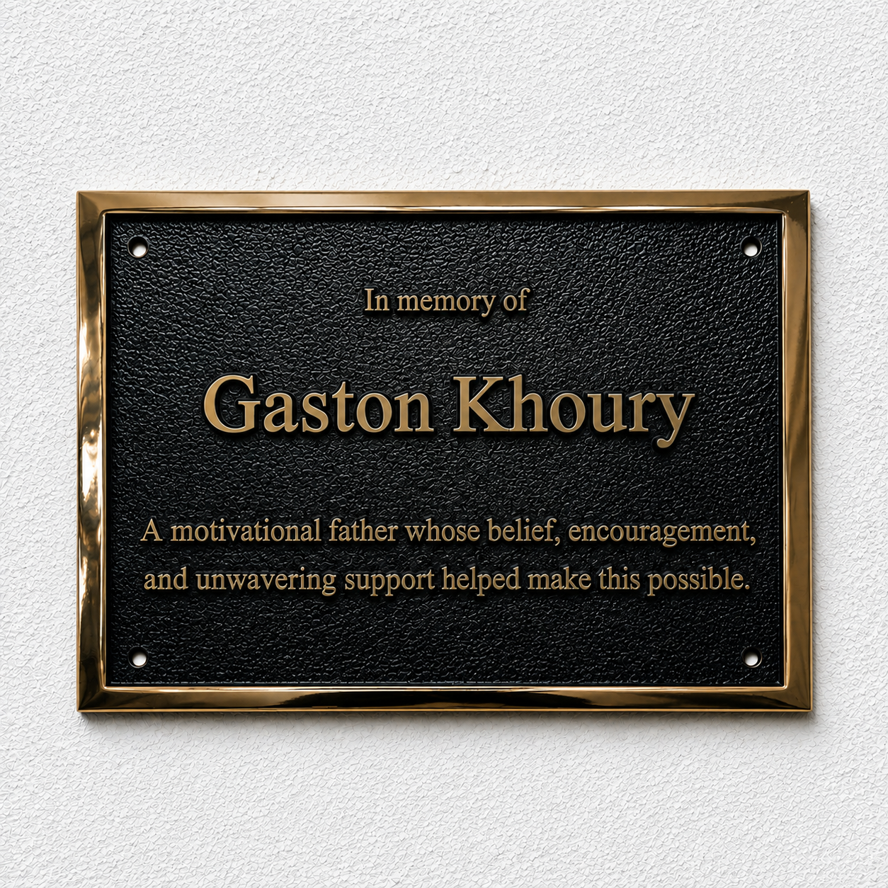 A rectangular black memorial plaque with a polished gold-toned border, mounted on a light textured wall. The plaque reads 'In memory of Gaston Khoury' and includes a short tribute below in gold lettering.