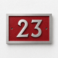 A modern house number plaque featuring the number 23 in raised silver-tone metal over a textured red background, mounted on a white textured wall.