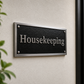 A rectangular black housekeeping sign with silver metallic lettering and border, mounted on a textured beige wall next to a dark door with a gold handle.