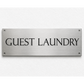 A rectangular brushed metal sign with the words 'GUEST LAUNDRY' in black serif lettering, mounted on a white textured wall.