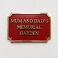 A rectangular red memorial garden sign with a gold border and raised gold lettering that reads “MUM AND DAD’S MEMORIAL GARDEN,” mounted on a textured white wall.