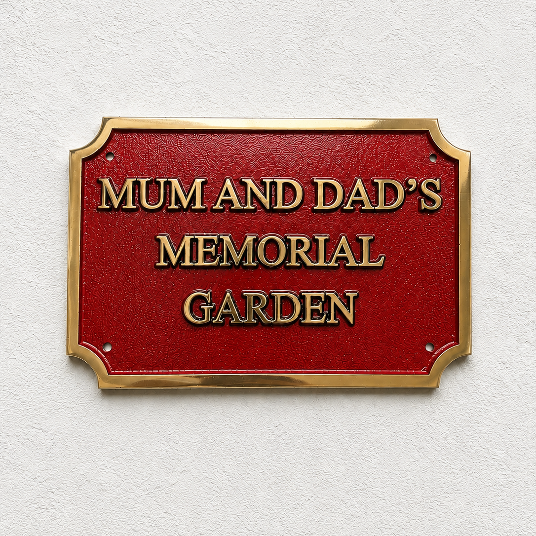 A rectangular red memorial garden sign with a gold border and raised gold lettering that reads “MUM AND DAD’S MEMORIAL GARDEN,” mounted on a textured white wall.