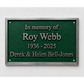 A rectangular green memorial plaque with a silver metal frame, mounted on a white textured wall. The plaque features raised silver text reading 'In memory of Roy Webb 1936 - 2025 Derek & Helen Bell-Jones'.