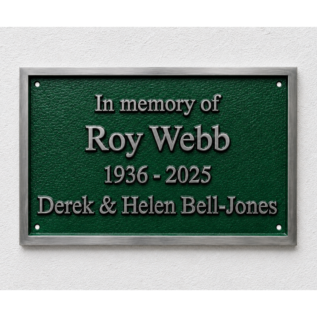 A rectangular green memorial plaque with a silver metal frame, mounted on a white textured wall. The plaque features raised silver text reading 'In memory of Roy Webb 1936 - 2025 Derek & Helen Bell-Jones'.