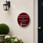 A round burgundy and white National Heritage Memorial Fund plaque mounted on a cream-colored stucco wall next to a black front door, with a black wall lantern and potted hydrangeas below.