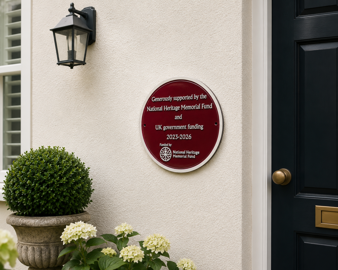 A round burgundy and white National Heritage Memorial Fund plaque mounted on a cream-colored stucco wall next to a black front door, with a black wall lantern and potted hydrangeas below.