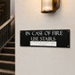 A black rectangular fire safety sign on a beige wall with the text 'IN CASE OF FIRE USE STAIRS' in white, alongside Braille text. To the left is a carpeted staircase with a dark metal railing and a lit wall sconce above.