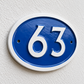 An oval blue and white house number plaque showing the number 63, mounted on a textured off-white wall.