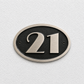 An oval house number plaque featuring the number 21 in brushed silver metal over a textured black center, mounted on a white textured wall.