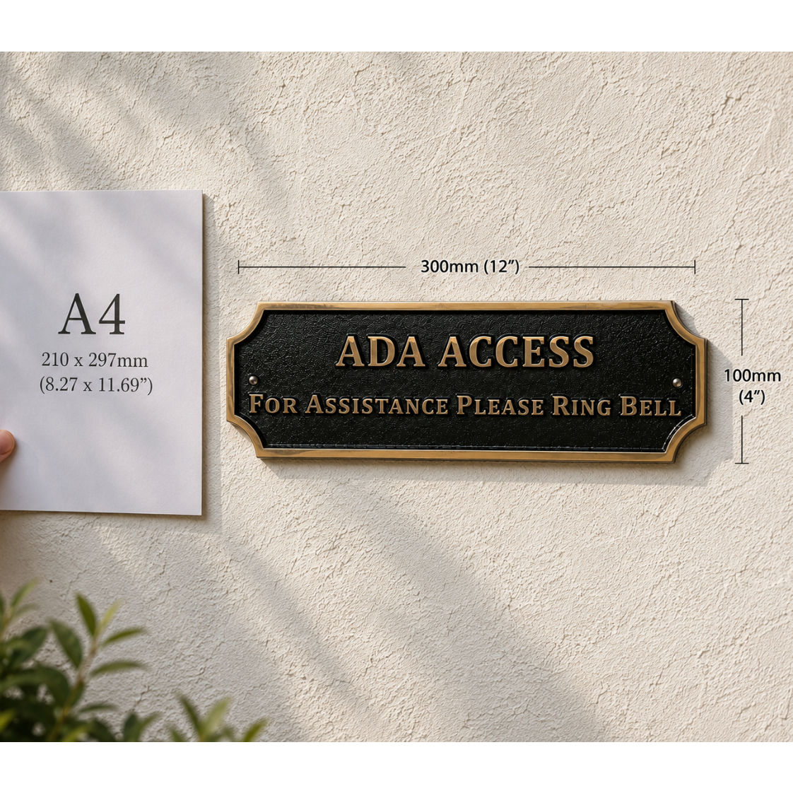 A black ADA Access plaque with gold lettering mounted on a textured beige wall, shown next to an A4 paper size reference held by a hand. The image includes dimension lines indicating the plaque is 300mm by 100mm.