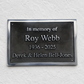 A rectangular dark metal memorial plaque mounted on a textured white wall, with raised silver text reading 'In memory of Roy Webb 1936 - 2025 Derek & Helen Bell-Jones'.