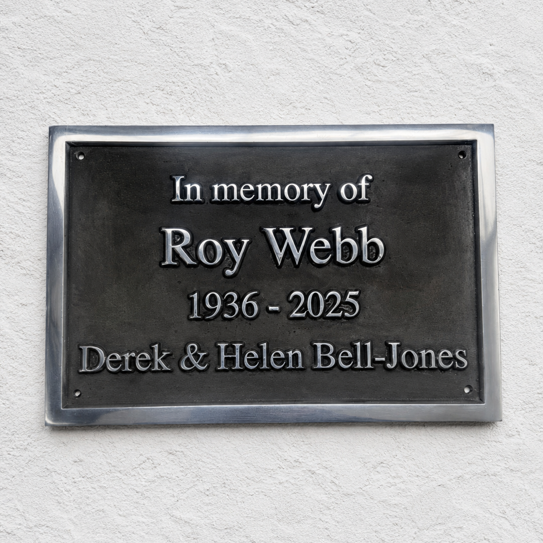 A rectangular dark metal memorial plaque mounted on a textured white wall, with raised silver text reading 'In memory of Roy Webb 1936 - 2025 Derek & Helen Bell-Jones'.