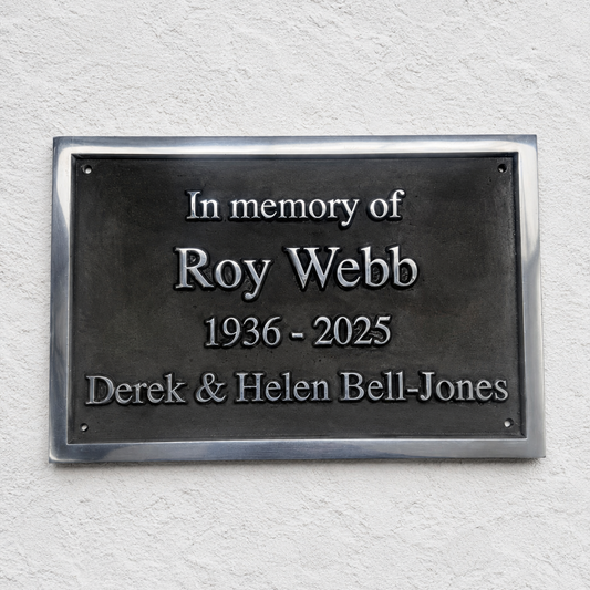 A rectangular dark metal memorial plaque mounted on a textured white wall, with raised silver text reading 'In memory of Roy Webb 1936 - 2025 Derek & Helen Bell-Jones'.