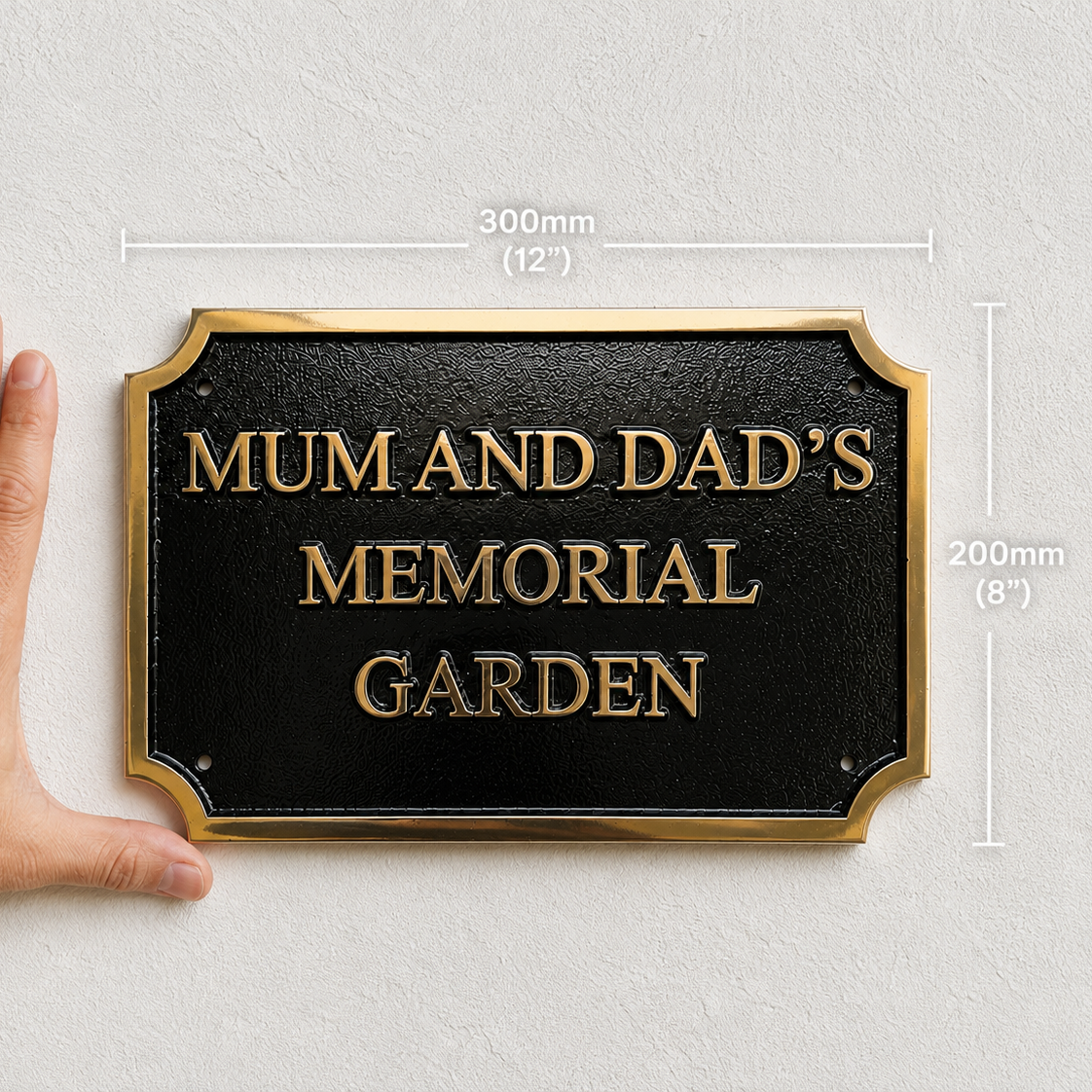 A black memorial garden plaque with gold lettering and a gold border, shown against a light textured wall with a hand beside it for scale. White measurement lines indicate 300mm by 200mm.