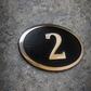 Cast bronze oval house number sign with polished raised numeral and black background
