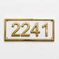 Gold house number plaque displaying '2241' in raised numerals on a white rectangular background, mounted on a textured white wall.
