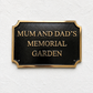 A rectangular black memorial garden sign with a gold border and raised gold lettering that reads “MUM AND DAD’S MEMORIAL GARDEN,” mounted on a textured white wall.