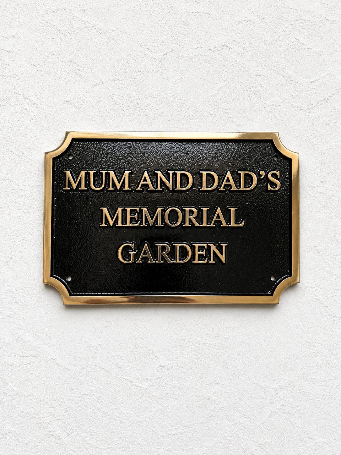 A rectangular black memorial garden sign with a gold border and raised gold lettering that reads “MUM AND DAD’S MEMORIAL GARDEN,” mounted on a textured white wall.