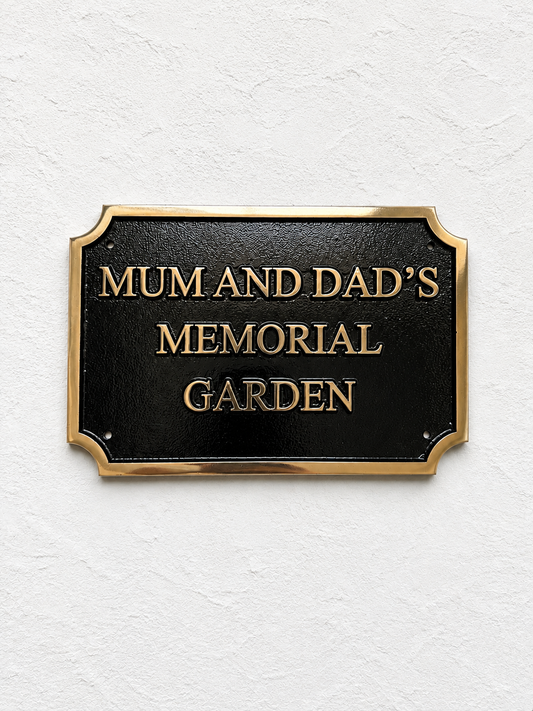 A rectangular black memorial garden sign with a gold border and raised gold lettering that reads “MUM AND DAD’S MEMORIAL GARDEN,” mounted on a textured white wall.