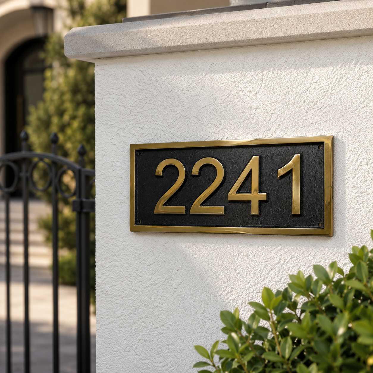 A rectangular black house number plaque with gold numerals and a gold border, mounted on a white stucco wall next to a black gate and green shrubs.