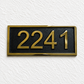 A rectangular house number plaque with the gold numerals '2241' on a black background, bordered by a gold frame, mounted on a textured white wall.