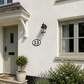 A white stucco house exterior with a pale green front door, a black wall-mounted lantern light, a circular house number 13 plaque, a potted topiary, and landscaped shrubs in the foreground.